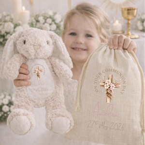 May include: A beige plush bunny and a beige drawstring fabric bag, both featuring a printed design of a wooden cross with pink flowers, the text "ON YOUR COMMUNION DAY", the name "Amelia", and the date "27th May 2026" in a script and sans-serif font.