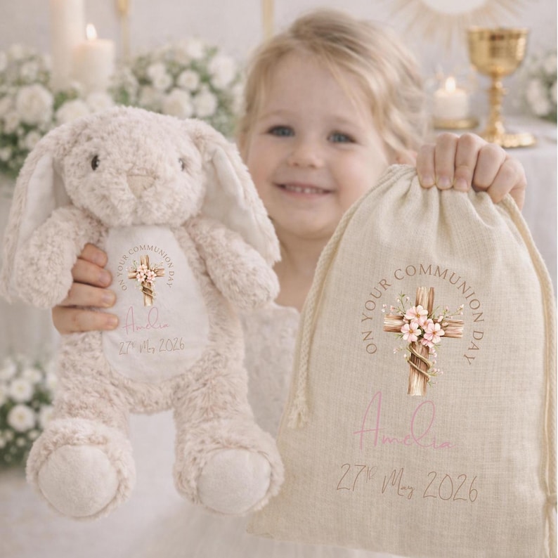 May include: A beige plush bunny and a beige drawstring fabric bag, both featuring a printed design of a wooden cross with pink flowers, the text "ON YOUR COMMUNION DAY", the name "Amelia", and the date "27th May 2026" in a script and sans-serif font.