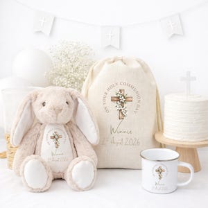 May include: A beige plush bunny, a drawstring linen bag, and a white enamel mug, all featuring a printed wooden cross with white flowers and the text "On Your Holy Communion Day," "Winnie," and "2nd August 2026" in a soft, neutral color palette.