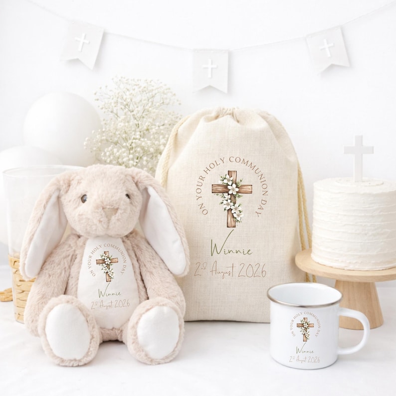 May include: A beige plush bunny, a drawstring linen bag, and a white enamel mug, all featuring a printed wooden cross with white flowers and the text "On Your Holy Communion Day," "Winnie," and "2nd August 2026" in a soft, neutral color palette.