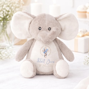 Op de afbeelding: Een zachtgrijze olifantenknuffel met grote oren en een slurf. De olifant heeft een witte borstvlek met de tekst "ON YOUR HOLY COMMUNION DAY" rond een kruis en bloemen. Daaronder staat de naam "Albert James" en de datum "18th July 2026".