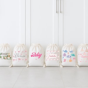 May include: Six off-white drawstring bags with various designs and names. One bag features jungle animals, another has pink flamingos, and a third displays the name "Betsy" in pink. Other bags have the names "Isla" and "Sophia" with colorful graphics.