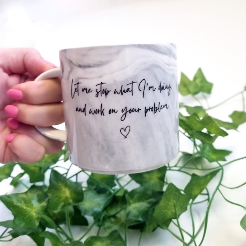 K&ouml;nnte beinhalten: Wei&szlig;e Keramiktasse mit Marmormuster und schwarzem Text, der "Lass mich aufh&ouml;ren, was ich tue, und an deinem Problem arbeiten." Ein kleines Herzsymbol befindet sich unter dem Text.
