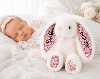 May include: A plush white stuffed rabbit toy featuring pink floral fabric on the inner ears and foot pads. The left ear is embroidered with the name Brooklynn, and the right ear displays the date 6-8-2026. A sheer pink ribbon is tied around its neck.