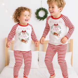May include: Two children wearing matching red and white striped Christmas pajamas. The long-sleeved shirts have a red collar and sleeves, and feature a wreath design with the text "The Christmas Tale of Mollie".