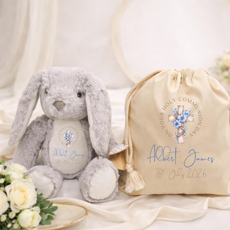 May include: A plush gray rabbit toy and a beige drawstring bag. Both items feature a printed design of a wooden cross adorned with blue flowers, the text "ON YOUR HOLY COMMUNION DAY", the name "Albert James", and the date "18th July 2026".
