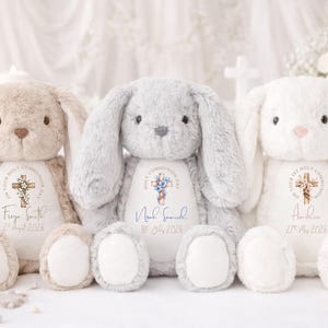 May include: Three plush bunny rabbits in beige, gray, and white. Each bunny has long ears and a soft, cuddly appearance. They feature a cross design with text, a name, and a date, likely for a religious occasion.
