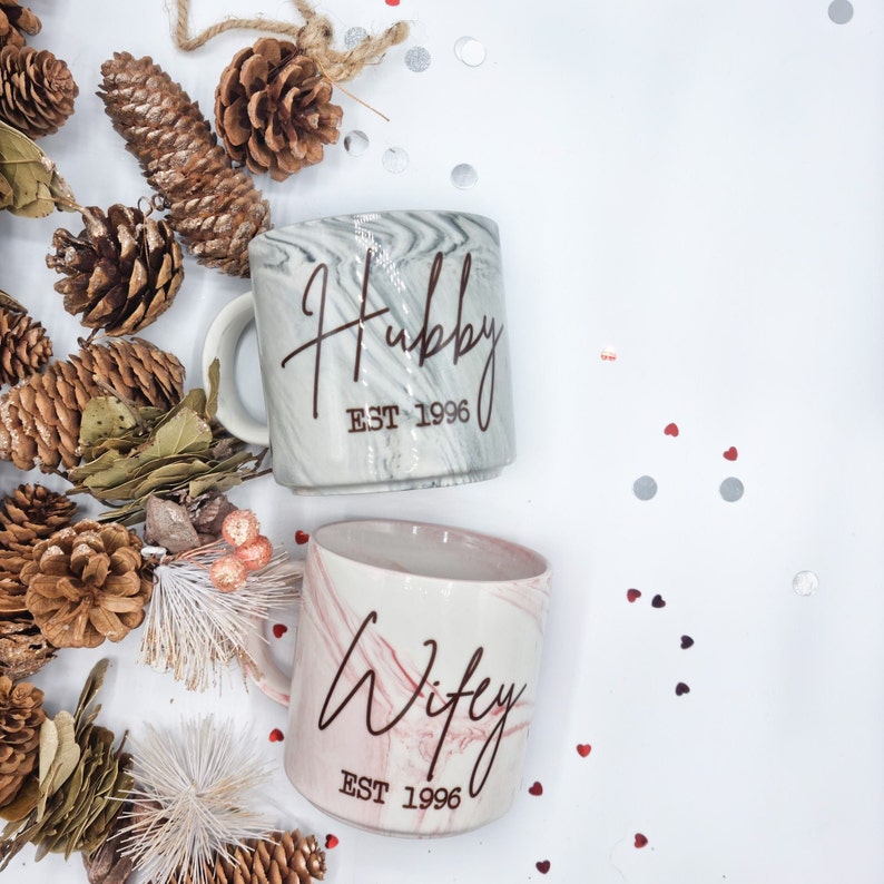 K&ouml;nnte beinhalten: Zwei Keramikbecher mit marmorierten Mustern. Ein Becher ist grau mit dem Text "Hubby EST 1996" und der andere ist rosa mit dem Text "Wifey EST 1996".