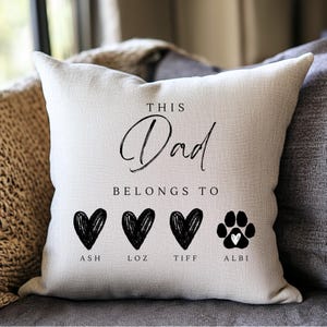May include: A cream-coloured cushion featuring the words "THIS Dad BELONGS TO" in black script. Below are three black hearts and a paw print with a heart, each with a name: Ash, Loz, Tiff, and Albi.