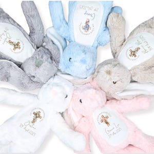 Op de afbeelding: Een verzameling pluche konijnen in pastelkleuren, waaronder grijs, blauw, wit en roze. Elk konijn heeft een gepersonaliseerde geborduurde patch met een kruis en tekst, inclusief namen en data, waarschijnlijk voor een doop.