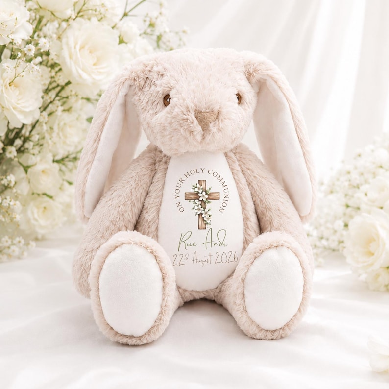 May include: A beige plush rabbit with long ears and a white belly patch. The belly features a printed wooden cross with white flowers, the text ON YOUR HOLY COMMUNION, the name Rue Andi, and the date 22nd August 2026 in a dark green font.