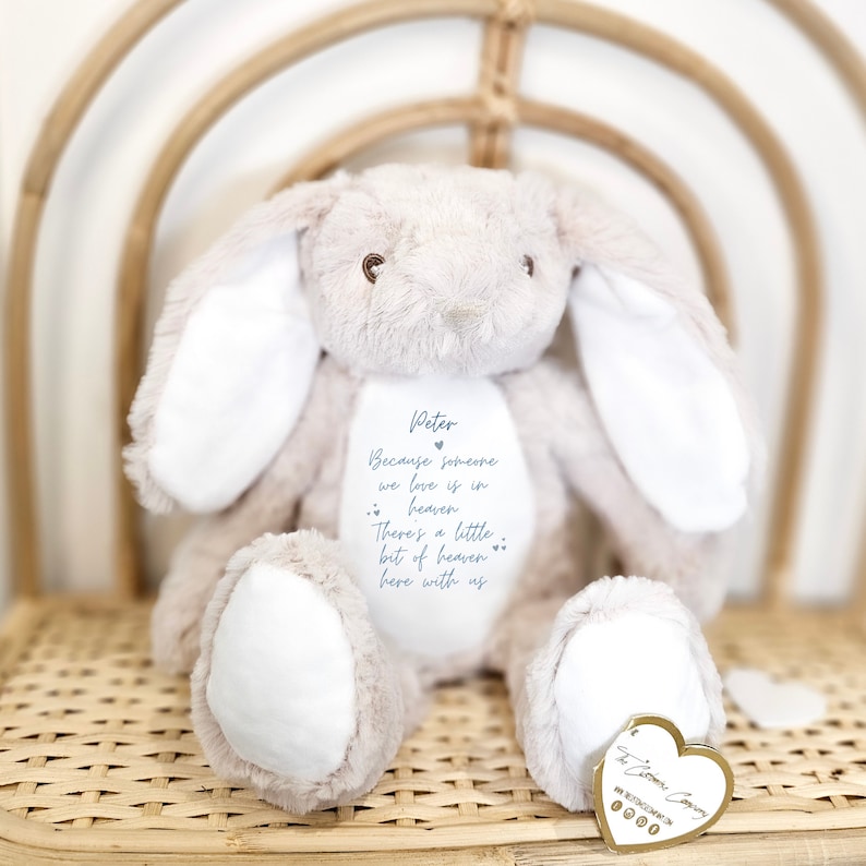 May include: A beige plush bunny with white ears and a heart-shaped tag. The bunny has a message embroidered on its chest that reads "Peter Because someone we love is in heaven There's a little bit of heaven here with us".