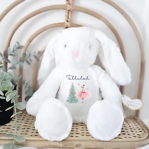 May include: A white plush bunny rabbit with long ears and a pink heart-shaped nose. The bunny has a graphic of a Christmas tree and a ballerina with the name "Tallulah" printed on its chest. The bunny is sitting on a woven wicker chair.