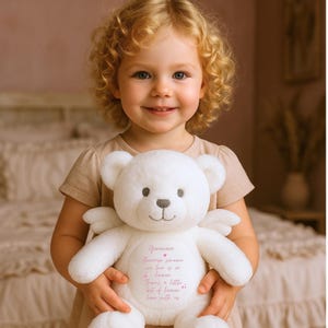 May include: A white teddy bear with angel wings holds a heart-shaped tag with the text "Genevieve Because someone we love is in heaven There's a little bit of heaven here with us".