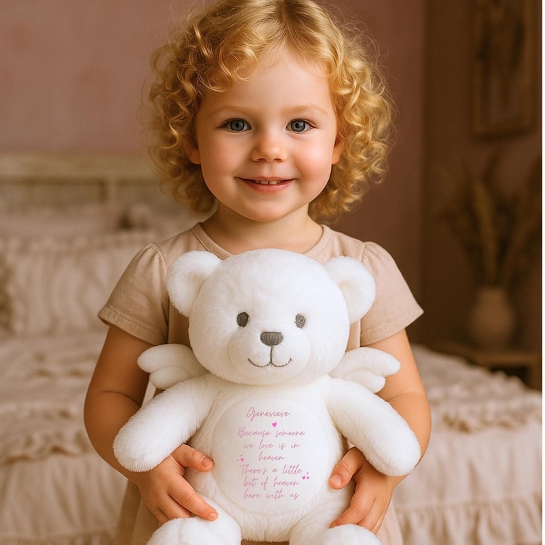May include: A white teddy bear with angel wings holds a heart-shaped tag with the text "Genevieve Because someone we love is in heaven There's a little bit of heaven here with us".