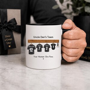 Op de afbeelding: Een witte keramische mok met een zwarte binnenkant en handvat, voorzien van een afbeelding van vier zwarte shirts aan een houten rek. De tekst luidt Uncle Dan's Team en Your Number One Fans, met namen en nummers op elk shirt gedrukt.