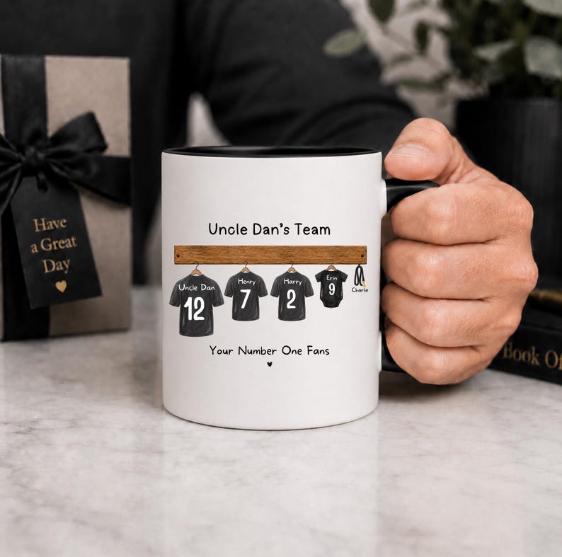 Op de afbeelding: Een witte keramische mok met een zwarte binnenkant en handvat, voorzien van een afbeelding van vier zwarte shirts aan een houten rek. De tekst luidt Uncle Dan's Team en Your Number One Fans, met namen en nummers op elk shirt gedrukt.