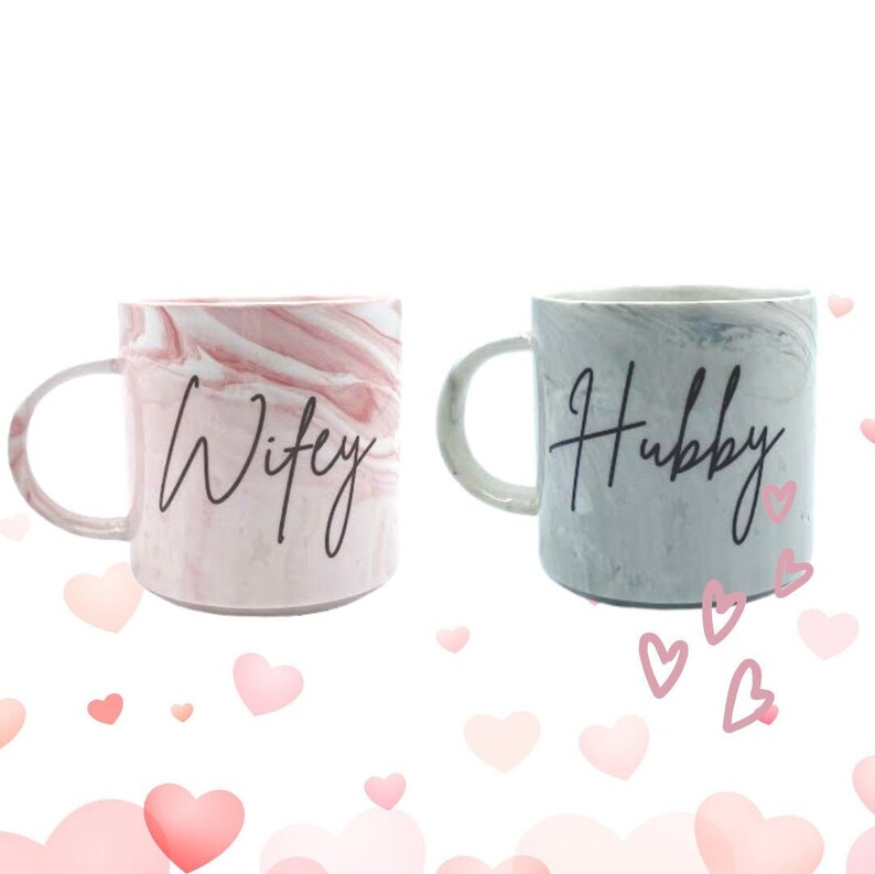 K&ouml;nnte beinhalten: Zwei Keramikbecher mit marmorierten Mustern. Der rosa Becher sagt "Wifey" und der graue Becher sagt "Hubby".