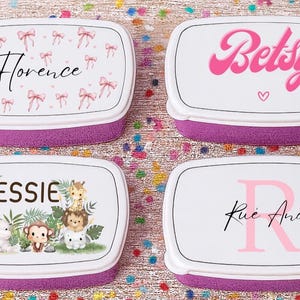May include: Four personalized lunch boxes with white lids and purple bases. The lids feature names and designs: "Florence" with bows, "Betsy" with a heart, "Jessie" with jungle animals, and "Rue Andi" with an initial.