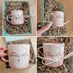 K&ouml;nnte beinhalten: Eine rosa-wei&szlig;e Marmortasse mit dem Text "Olivia's Mum" in schwarzer Schrift und einem kleinen Herz. Die Tasse befindet sich in einer Geschenkbox mit zerkn&uuml;lltem Papier.