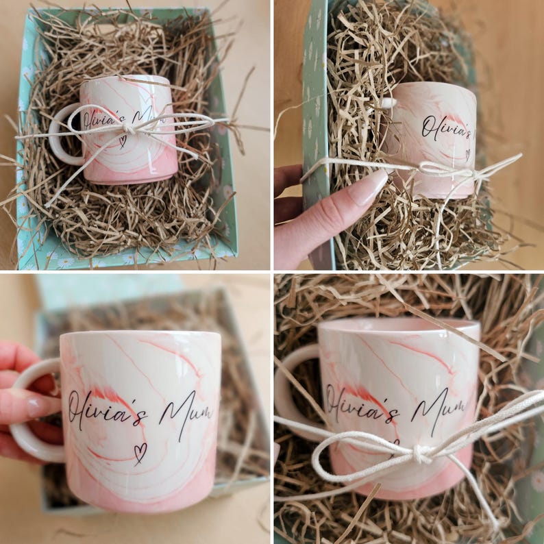 K&ouml;nnte beinhalten: Eine rosa-wei&szlig;e Marmortasse mit dem Text "Olivia's Mum" in schwarzer Schrift und einem kleinen Herz. Die Tasse befindet sich in einer Geschenkbox mit zerkn&uuml;lltem Papier.