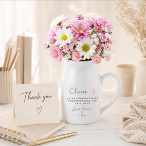 May include: A white ceramic pitcher mug filled with a bouquet of pink and white daisies. The mug features the text: Claire, NEVER UNDERESTIMATE THE DIFFERENCE YOU HAVE MADE, Love from ISLA. A card reads: Thank you, with a gold heart symbol.