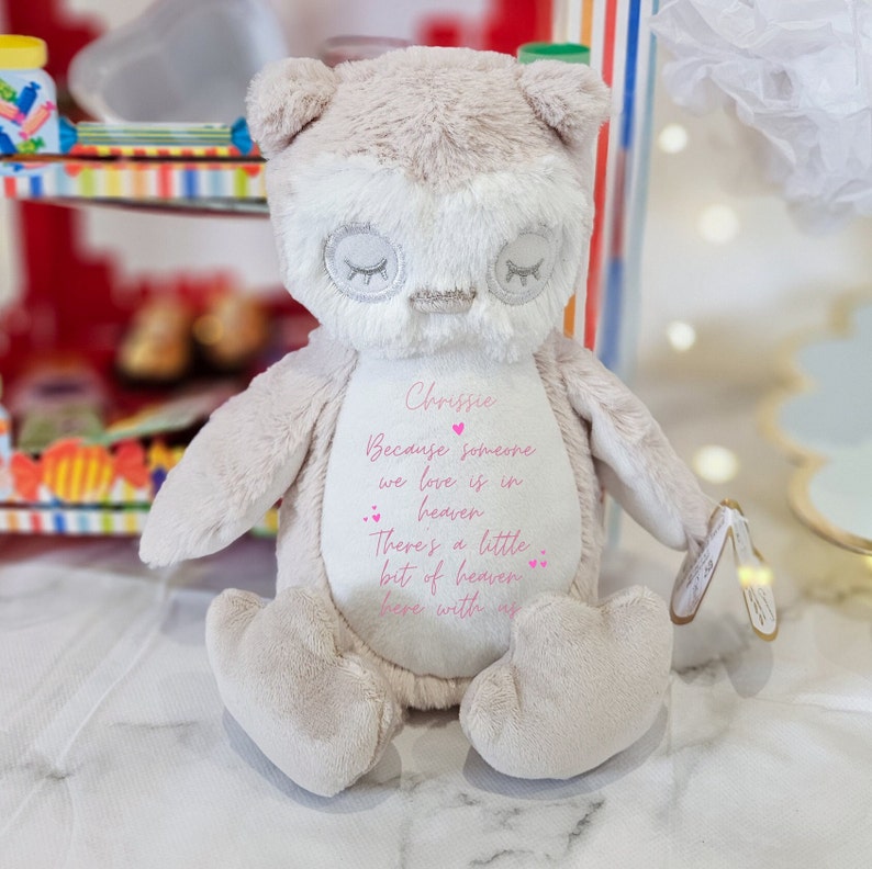 May include: A plush owl toy with a beige and white color scheme. The owl has embroidered eyes and a pink heart on its chest. The owl is sitting on a white surface and has the text "Chrissie Because someone we love is in heaven There's a little bit of heaven here with us" embroidered on its chest.
