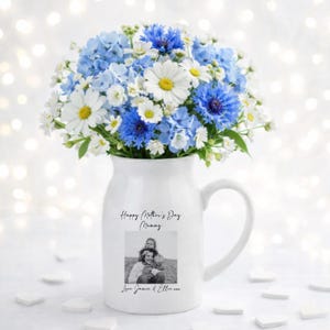 May include: A white ceramic pitcher with a handle, filled with a bouquet of blue and white flowers. The pitcher has a black and white photo and text that reads "Happy Mother's Day Mummy" and "Love Jamie & Ellie".