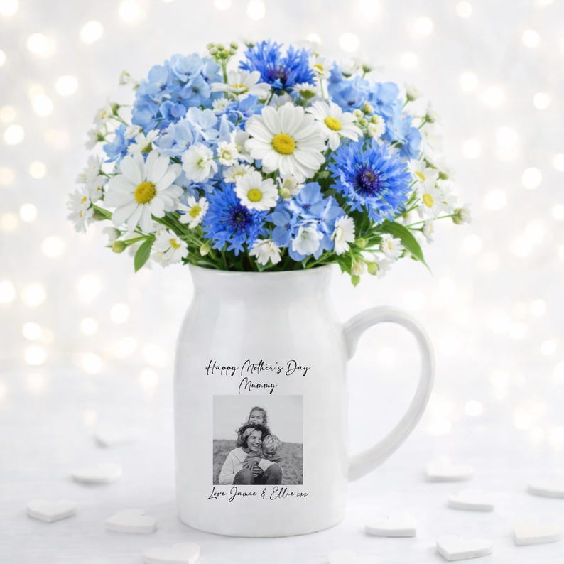 May include: A white ceramic pitcher with a handle, filled with a bouquet of blue and white flowers. The pitcher has a black and white photo and text that reads "Happy Mother's Day Mummy" and "Love Jamie & Ellie".