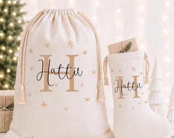 Pode incluir: Um saco de presente cor de creme com cordão e uma meia de Natal a condizer, ambos com o nome Hattie em letra cursiva preta sobre uma letra H grande em tom castanho claro, rodeados por estrelas. Os artigos têm cordões de corda entrançada.