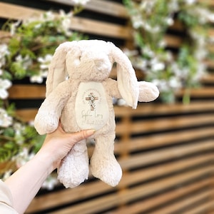 Op de afbeelding: Een zacht, beige pluche konijn met lange oren. Het konijn heeft een label op de borst met de tekst "ON YOUR CHRISTENING DAY" en een kruisontwerp. Daaronder staat de naam "Sophia Long" en de datum "2nd August 2025".