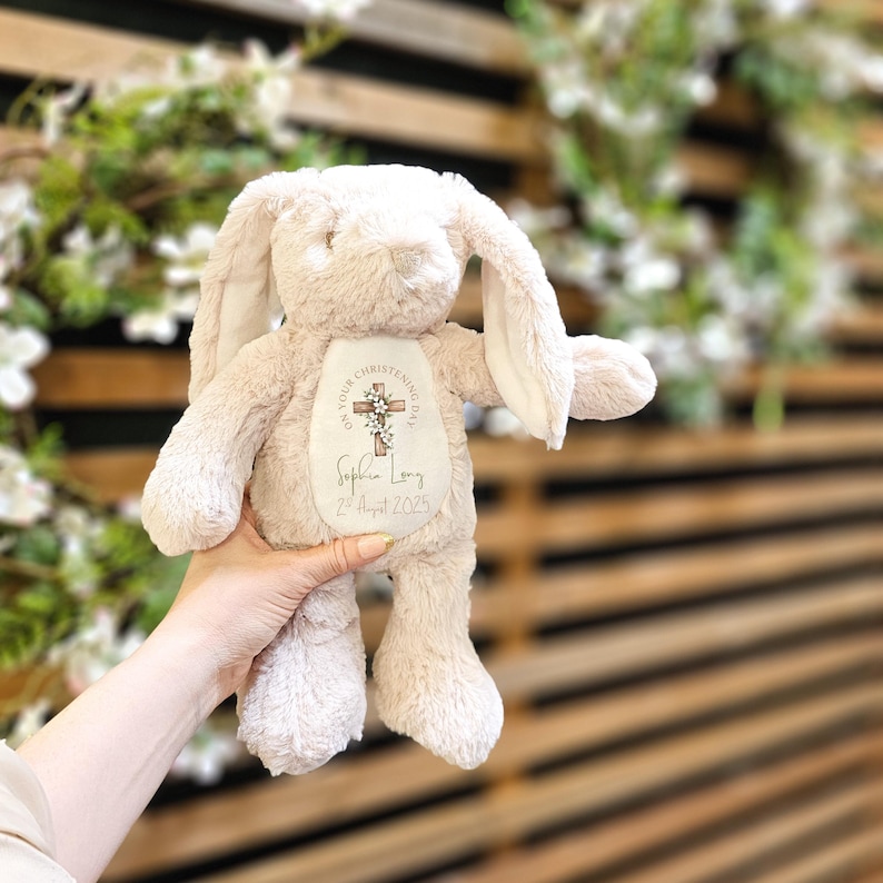 Op de afbeelding: Een zacht, beige pluche konijn met lange oren. Het konijn heeft een label op de borst met de tekst "ON YOUR CHRISTENING DAY" en een kruisontwerp. Daaronder staat de naam "Sophia Long" en de datum "2nd August 2025".