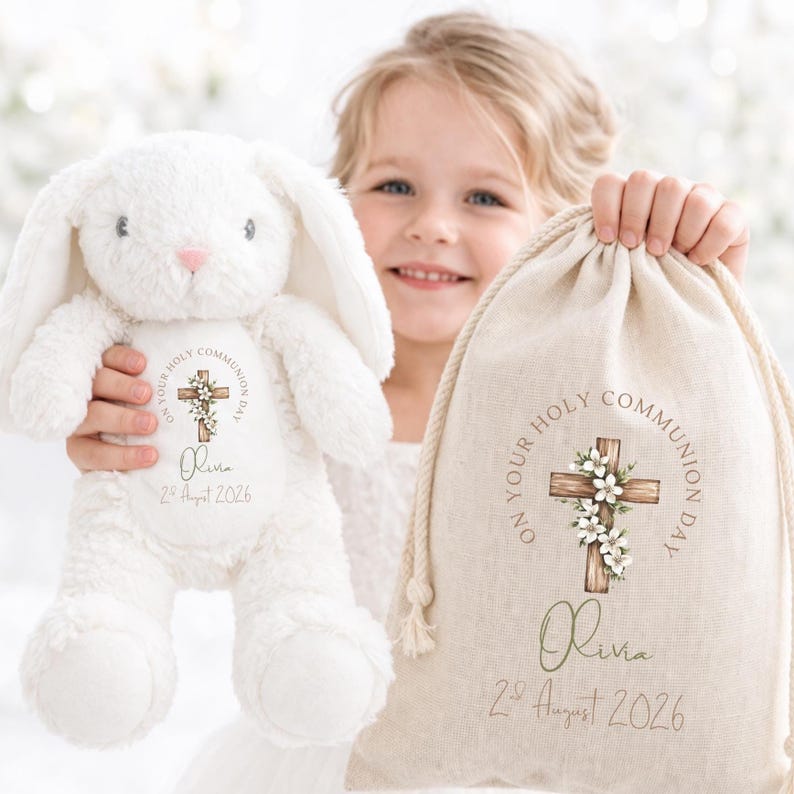 Op de afbeelding: Een witte pluche konijn en een beige trekkoordtas. Beide items zijn gepersonaliseerd met de tekst "ON YOUR HOLY COMMUNION DAY", een kruis met bloemaccenten, de naam "Olivia" en de datum "2 augustus 2026".