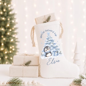 Puede incluir: Un calcetín navideño de tela blanca con cierre de cordón, que presenta una ilustración impresa de un pingüino junto a un árbol de Navidad azul y una caja de regalo. El texto "Special Delivery For" y el nombre "Elsie" están impresos en azul.