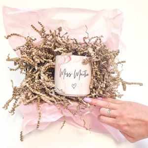 K&ouml;nnte beinhalten: Eine rosa und wei&szlig;e marmorierte Keramiktasse mit dem Text "Miss Martha" und einem kleinen Herzdesign. Die Tasse befindet sich in einer rosa Geschenkpapier-Tasche, gef&uuml;llt mit braunem Seidenpapier.