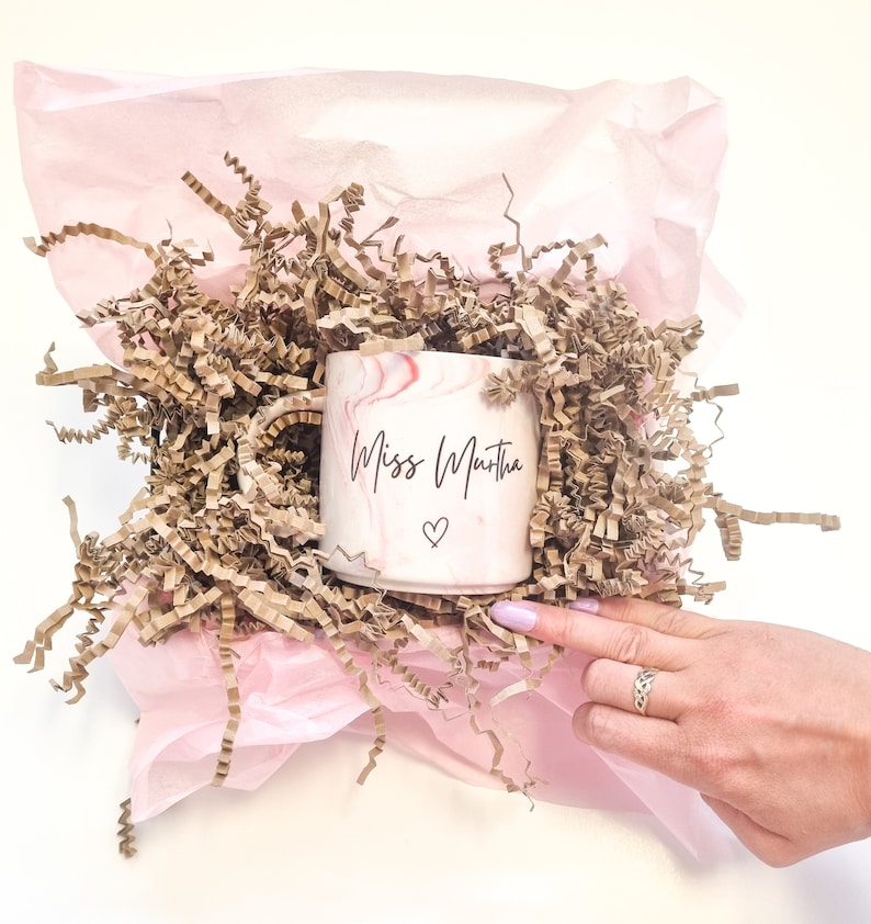 K&ouml;nnte beinhalten: Eine rosa und wei&szlig;e marmorierte Keramiktasse mit dem Text "Miss Martha" und einem kleinen Herzdesign. Die Tasse befindet sich in einer rosa Geschenkpapier-Tasche, gef&uuml;llt mit braunem Seidenpapier.