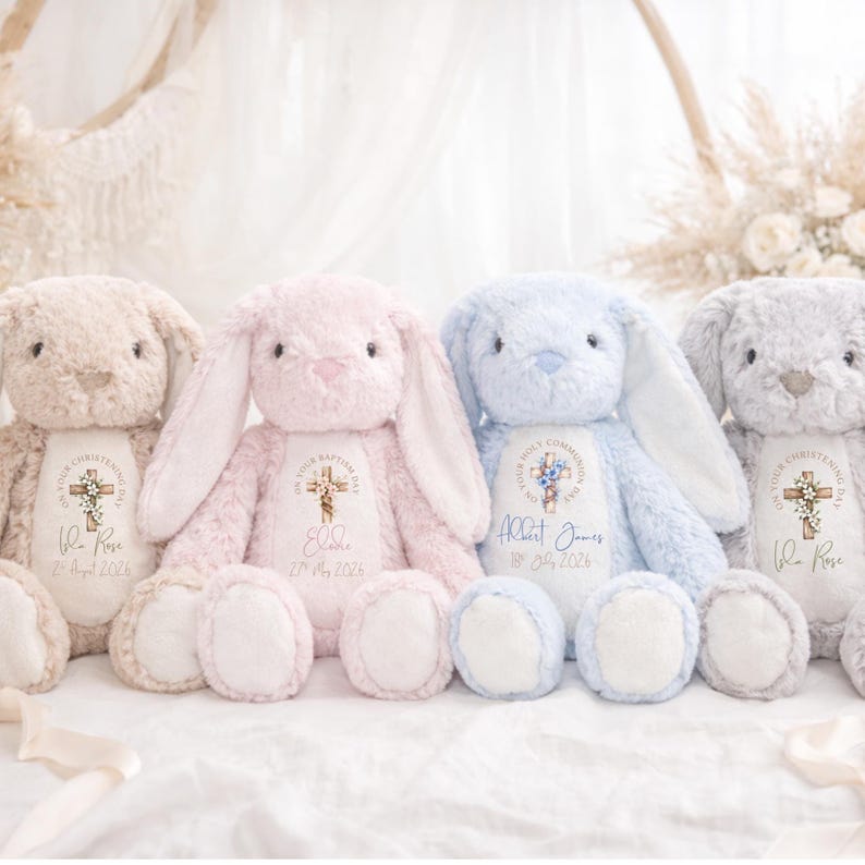 Op de afbeelding: Vier pluche konijnen in beige, roze, blauw en grijs. Elk konijn heeft lange oren, een rond lichaam en een gepersonaliseerde geborduurde patch met een kruis en tekst. De konijnen zitten op een wit oppervlak.
