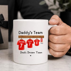 Op de afbeelding: Een witte keramische mok met de tekst Daddy's Team en Dad's Dream Team. Het ontwerp toont drie rode sportshirts aan een houten rek, gelabeld Daddy 7, Sophie 7 en Coby 9, samen met een rode hondenriem met de naam Maxi en een klein hartje.