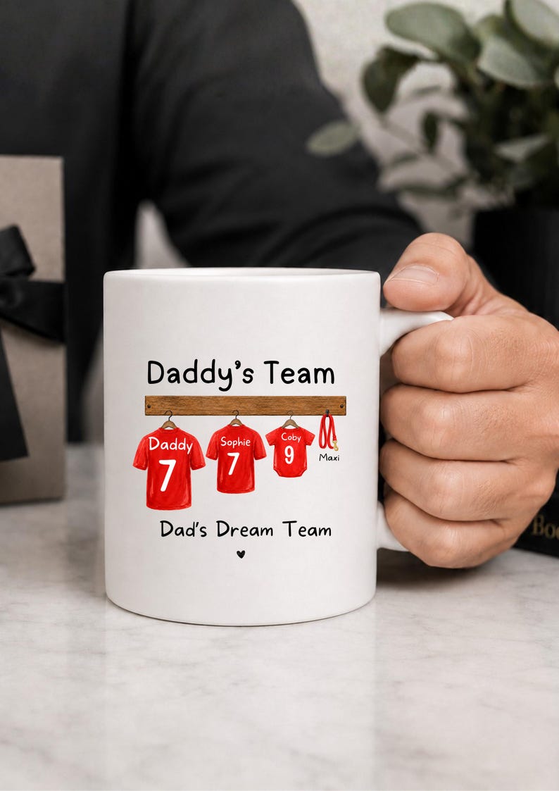 Op de afbeelding: Een witte keramische mok met de tekst Daddy's Team en Dad's Dream Team. Het ontwerp toont drie rode sportshirts aan een houten rek, gelabeld Daddy 7, Sophie 7 en Coby 9, samen met een rode hondenriem met de naam Maxi en een klein hartje.