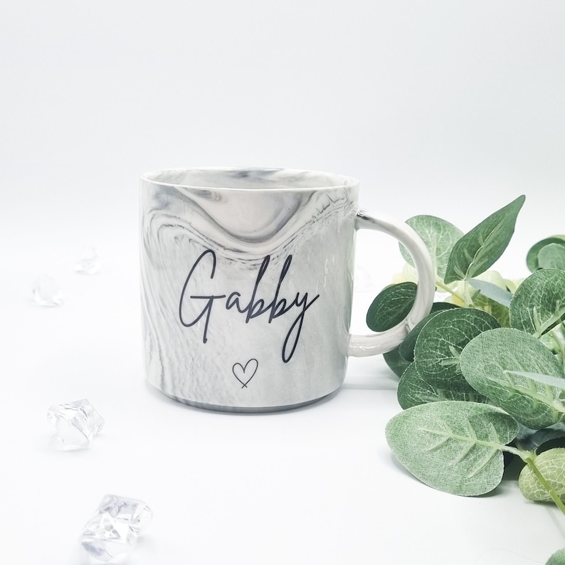 May include: A white ceramic mug with a gray marble pattern and the name "Gabby" written in black script. A small heart is drawn below the name.