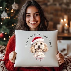 May include: A square, off-white pillow featuring a watercolor illustration of a dog wearing a Santa hat. The text "HAVE A MAGICAL CHRISTMAS" arches above the dog, with the name "Freya" below. The dog has light brown fur and a happy expression.