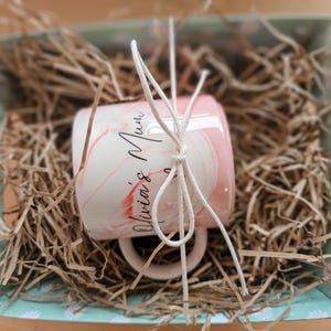 K&ouml;nnte beinhalten: Eine rosa-wei&szlig;e Keramiktasse mit dem Text "Olivia's Mum" in schwarzer Kalligraphie. Die Tasse ist mit wei&szlig;em Garn umwickelt und in einer hellblauen und wei&szlig;en Geschenkbox mit braunem, zerkleinertem Papier verpackt.