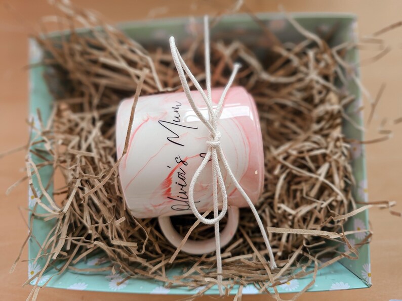 K&ouml;nnte beinhalten: Eine rosa-wei&szlig;e Keramiktasse mit dem Text "Olivia's Mum" in schwarzer Kalligraphie. Die Tasse ist mit wei&szlig;em Garn umwickelt und in einer hellblauen und wei&szlig;en Geschenkbox mit braunem, zerkleinertem Papier verpackt.