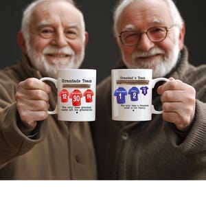 Op de afbeelding: Twee witte keramische mokken met bedrukte afbeeldingen van hangende sportshirts. De linker mok toont rode shirts met de nummers 12, 30, 14 en de tekst Grandads Team. De rechter mok toont blauwe shirts met de nummers 1, 2, 3 en de tekst.