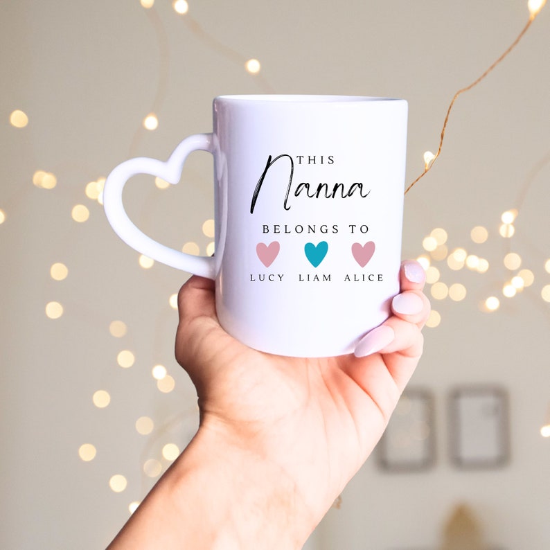May include: White ceramic mug with a heart-shaped handle. The mug has black text that reads "This Nanna belongs to" with three hearts below, labeled "Lucy", "Liam", and "Alice".