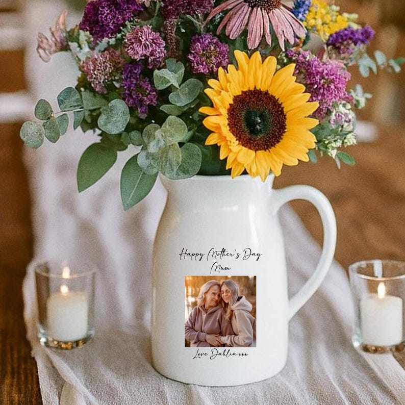 May include: A white ceramic pitcher with a handle, filled with a vibrant bouquet of flowers, including a large yellow sunflower. The pitcher has a photo and text that reads "Happy Mother's Day Mum". Two lit candles are in glass holders.