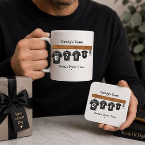 Op de afbeelding: Een witte keramische mok en een bijpassende vierkante onderzetter, beide voorzien van een afbeelding van vier zwarte sportshirts aan een houten rek met de tekst Daddy's Team en Daddy's Dream Team, naast een geschenkdoos met zwart lint.