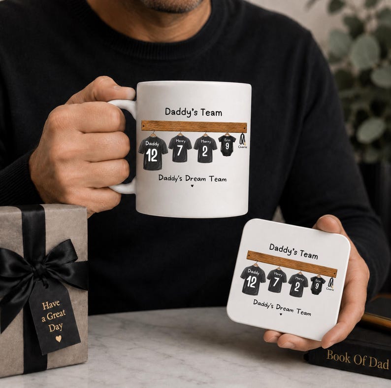Op de afbeelding: Een witte keramische mok en een bijpassende vierkante onderzetter, beide voorzien van een afbeelding van vier zwarte sportshirts aan een houten rek met de tekst Daddy's Team en Daddy's Dream Team, naast een geschenkdoos met zwart lint.