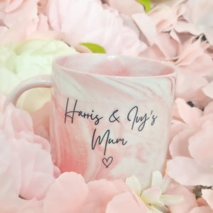 K&ouml;nnte beinhalten: Eine rosa-wei&szlig;e Marmortasse mit dem Text "Harris & Ivy's Mum" und einem Herzsymbol. Die Tasse ist von rosa Bl&uuml;tenbl&auml;ttern umgeben.