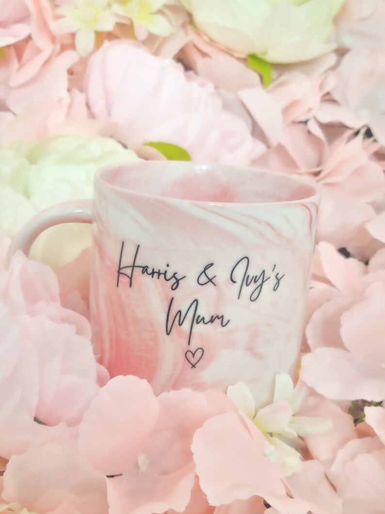 K&ouml;nnte beinhalten: Eine rosa-wei&szlig;e Marmortasse mit dem Text "Harris & Ivy's Mum" und einem Herzsymbol. Die Tasse ist von rosa Bl&uuml;tenbl&auml;ttern umgeben.